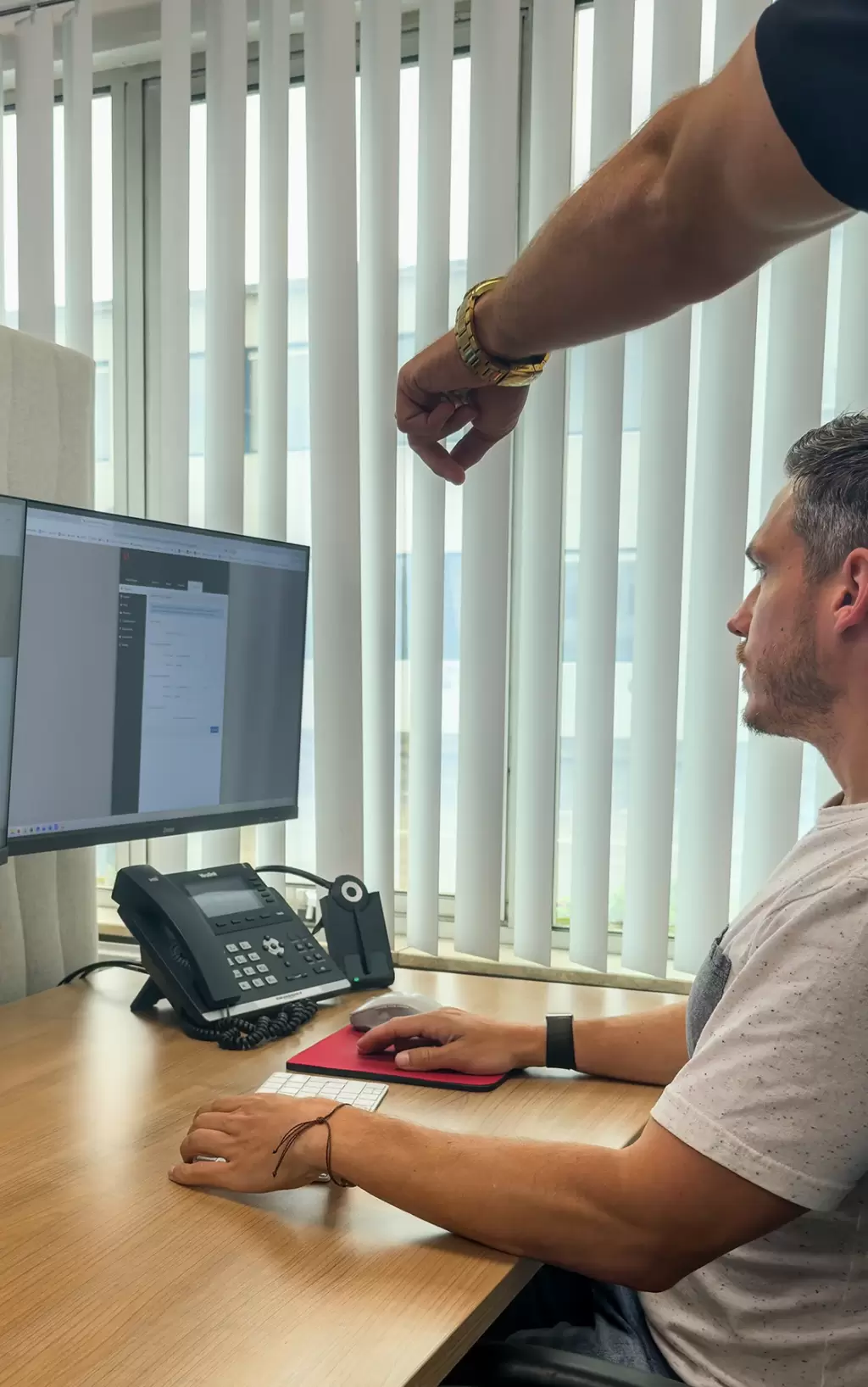 BCSA-engineer aan zijn werkplek met dubbel scherm en een Yealink-toestel, terwijl een collega meekijkt.